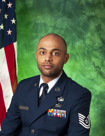 TSgt Fabio Deavers posing in front of a green background and U.S. flag. 