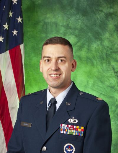 Major Paul Frisinger posing in front of green background and U.S. flag. 